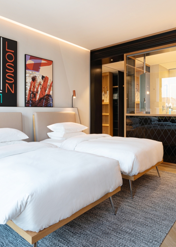 Elegantes Hotelzimmer im modernen Design mit zwei Betten und offenes Badezimmer hinter einer Glasscheibe.