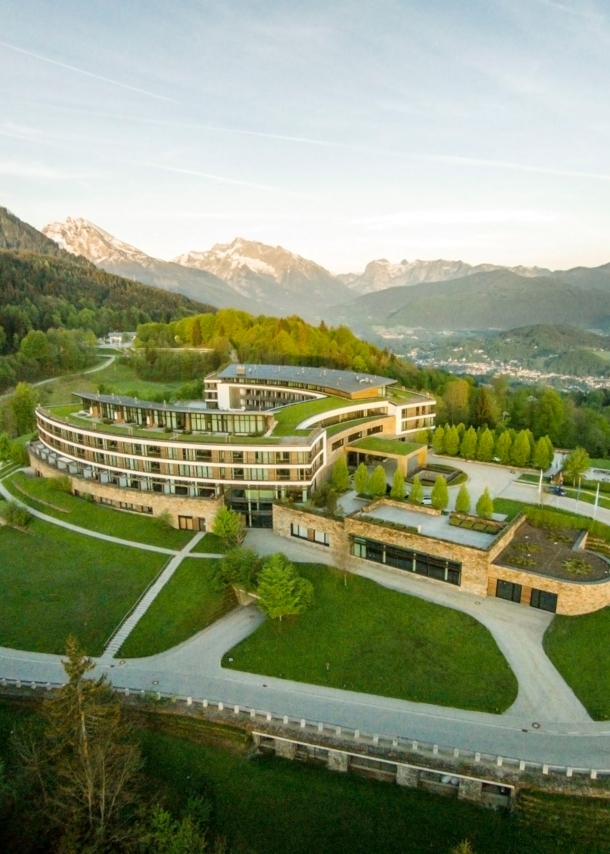 Luftaufnahme eines hufeisenförmigen Hotelkomplexes mit Rasenflächen inmitten einer hügeligen Waldlandschaft.