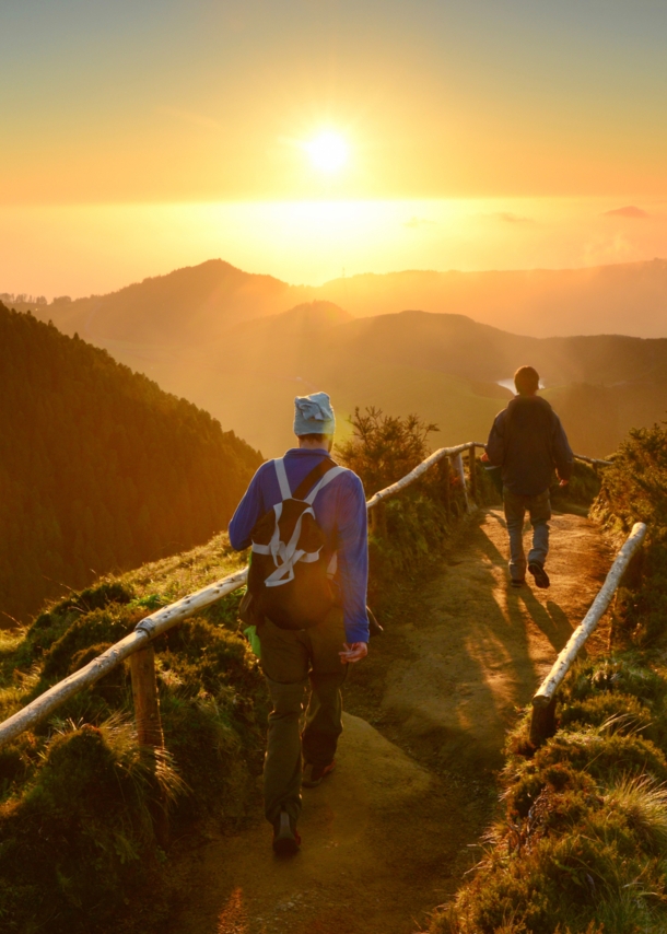 Rückansicht zweier gehender Personen auf einem Weg entlang eines Bergkamms inmitten einer grünen Gebirgslandschaft bei Sonnenuntergang.