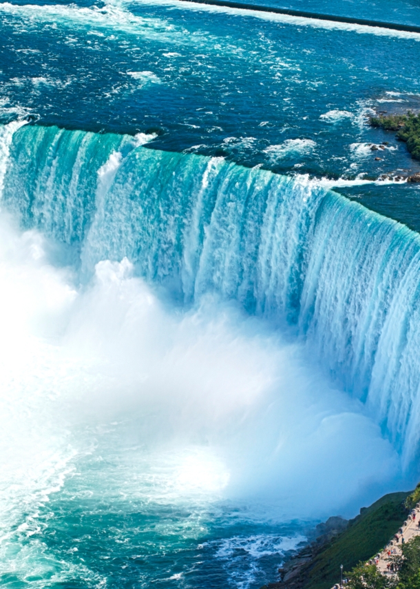 Luftaufnahme eines runden Wasserbeckens mit Boot vor einem Wasserfall, am Ufer eine Aussichtsplattform mit Personen.