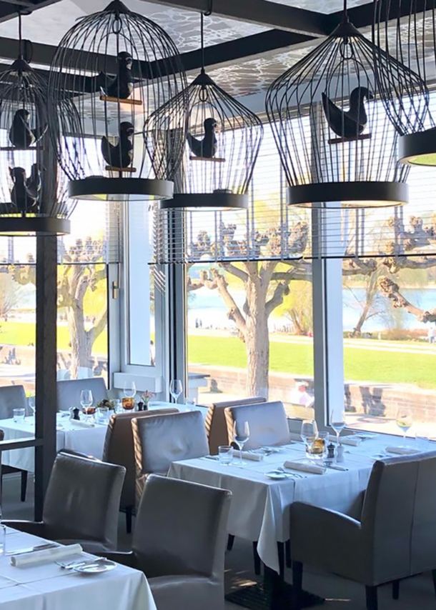 Speiseraum des Restaurants OX Royal mit eingedeckten Tischen und dekorativen Vogelkäfigen an der Decke in einem Wintergarten mit Panoramablick an der Uferpromenade eines Flusses.