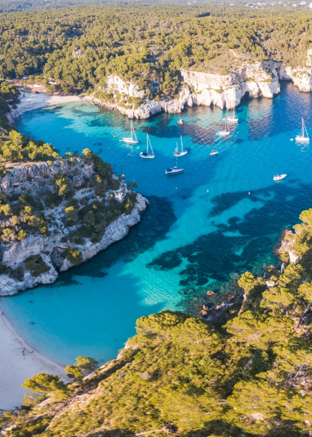 Luftaufnahme eines Küstenabschnitts von Menorca mit Badebuchten, Waldlandschaft und Booten.