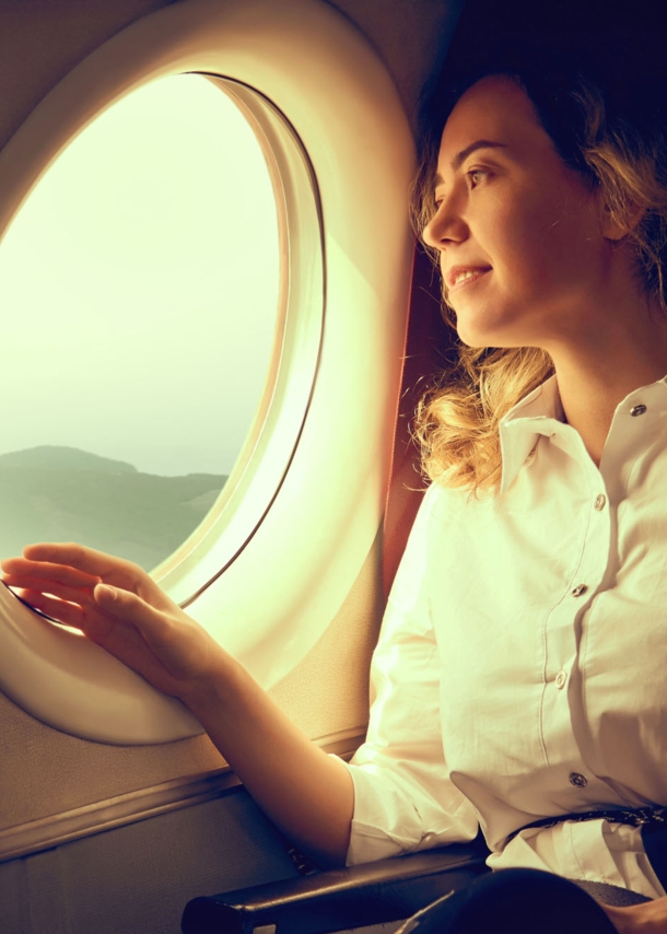 Eine junge Frau in weißer Bluse schaut aus dem Fenster eines Flugzeuges auf eine Landschaft.