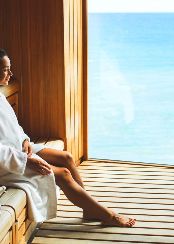 Eine junge Frau im weißen Bademantel sitzt entspannt in einer Holzsauna mit Panoramafenster am Meer.