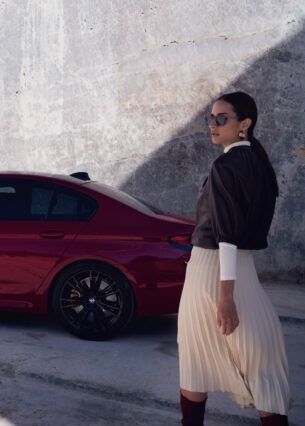 Eine elegante Frau mit Sonnenbrille vor einem roten BMW vor einer Betonwand.