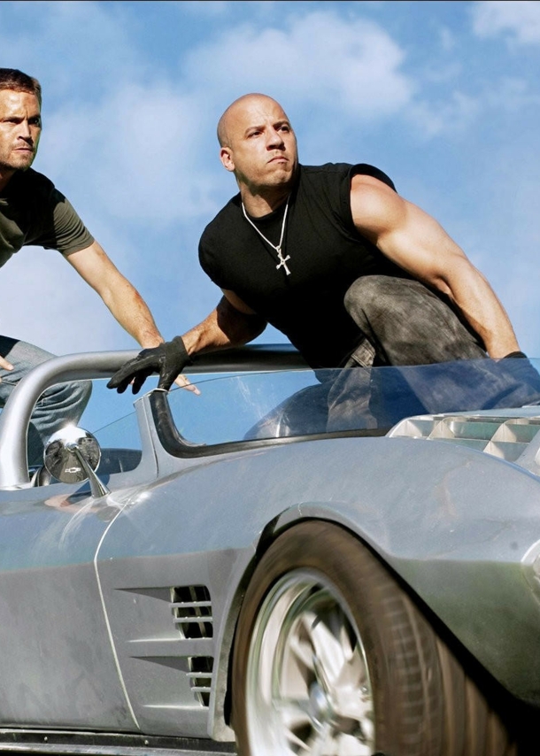 Paul Walker und Vin Diesel hocken auf einem silbernen Sportwagen ohne Verdeck.