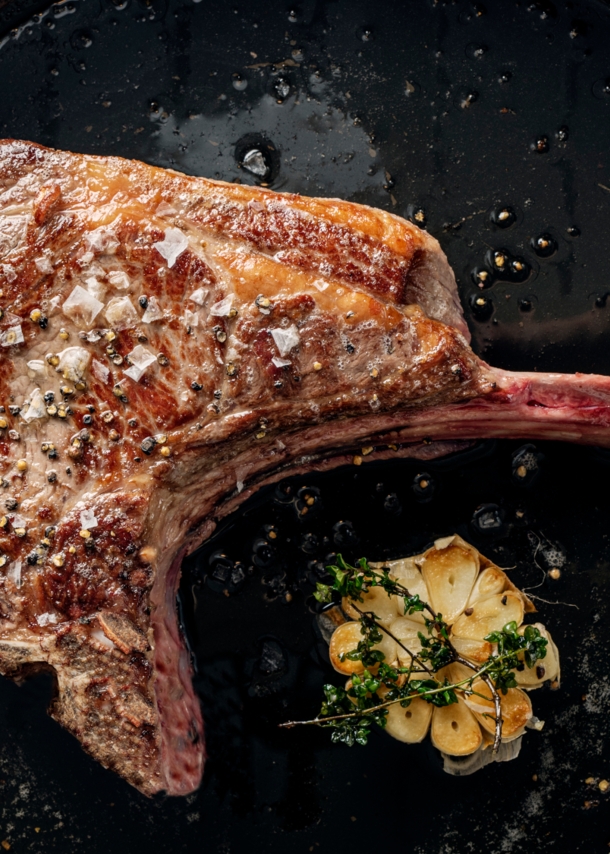 Ein scharf angebratenes Tomahawk-Steak in einer Pfanne. Daneben liegt eine aufgeschnittene Knoblauchknolle, die mit Thymian getoppt ist.