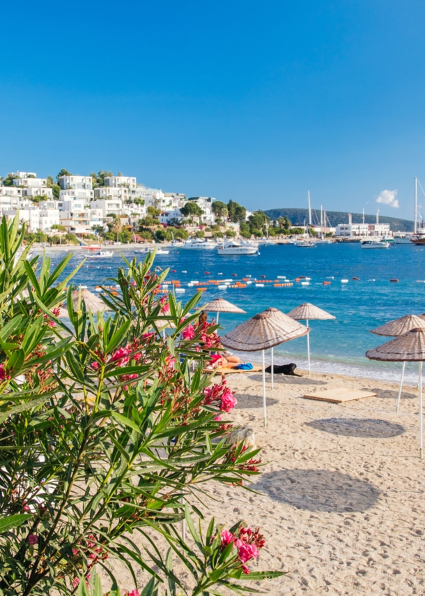 Strand in Bodrum mit Sonnenschirmen mit weißen Häusern an der Küste im Hintergrund.