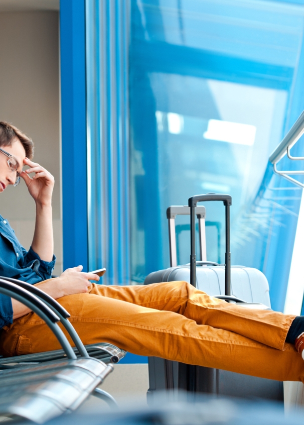 Ein Mann, der am Flughafen auf einer Bankreihe mit ausgestreckten Beinen sitzt. In einer Hand hält er ein Smartphone, die andere Hand ist an seiner Schläfe. Neben ihm steht ein Koffer.