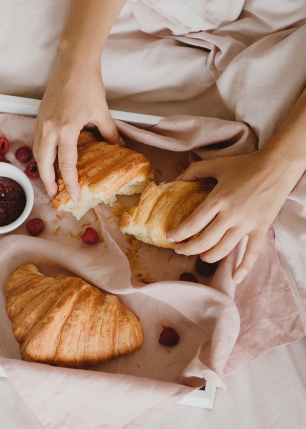 Auf einem Bett liegt ein Tablett mit Croissants, einer Tasse Kaffee, ein Schälchen Marmelade, einem Löffel und Früchten. Dahinter sitzt eine unkenntliche Person, die eines der Croissants auseinanderreißt.