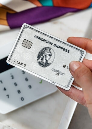 Nahaufnahme einer Hand, die eine silberne American Express Kreditkarte an ein Kartenlesegerät neben Modeaccessoires hält.