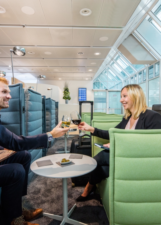 Ein Mann und eine Frau sitzen sich gegenüber auf Sesseln in einer Flughafenlounge und stoßen mit Weingläsern an.