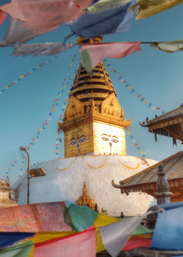 Goldener Turm einer buddhistische Tempelanlage mit aufgemalten Gesichtern neben einer Pagode, im Vordergrund wehende, bunte Gebetsflaggen.