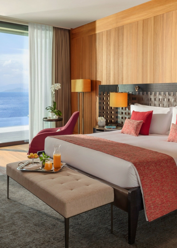 Modern eingerichtete Suite des Hotels Mandarin Oriental Bodrum mit einem großen Bett, einer Sitzbank, Arbeitstisch und raumhohen Fenstern mit Meeresblick.