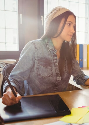 Frau sitzt an ihrem Schreibtisch und arbeitet am Computer sowie am Tablet.