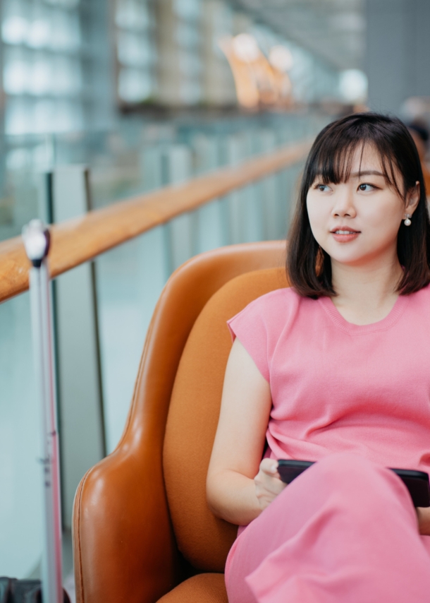 Eine asiatische Frau in einem pinken Kleid sitzt in einem braunen Ledersessel auf einer Galerie einer Flughafenlounge.