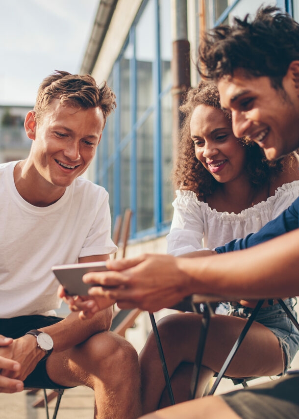 Eine Gruppe von Menschen sitzt zusammen auf dem Balkon und schaut gemeinsam etwas auf dem Smartphone.