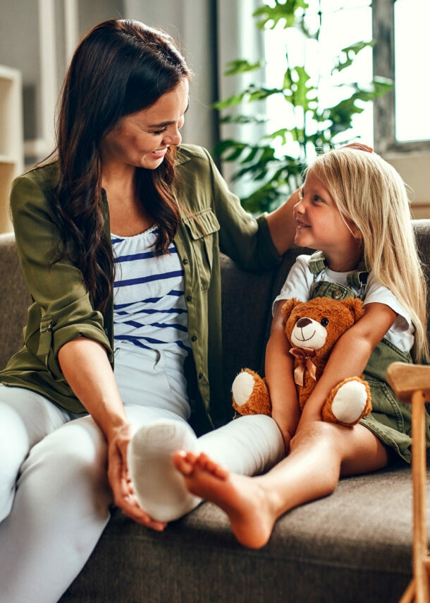 Ein kleines Mädchen mit eingegipstem Bein und Teddybär auf dem Schoß wird von ihrer Mutter auf dem Sofa getröstet.