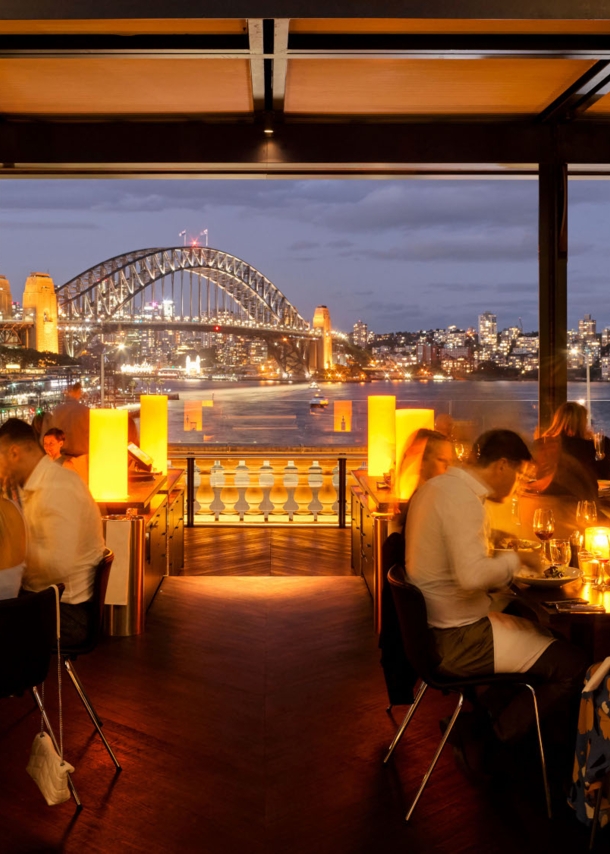 Außengastronomie am Hafen von Sydney vor beleuchteter Harbour Bridge bei Nacht.