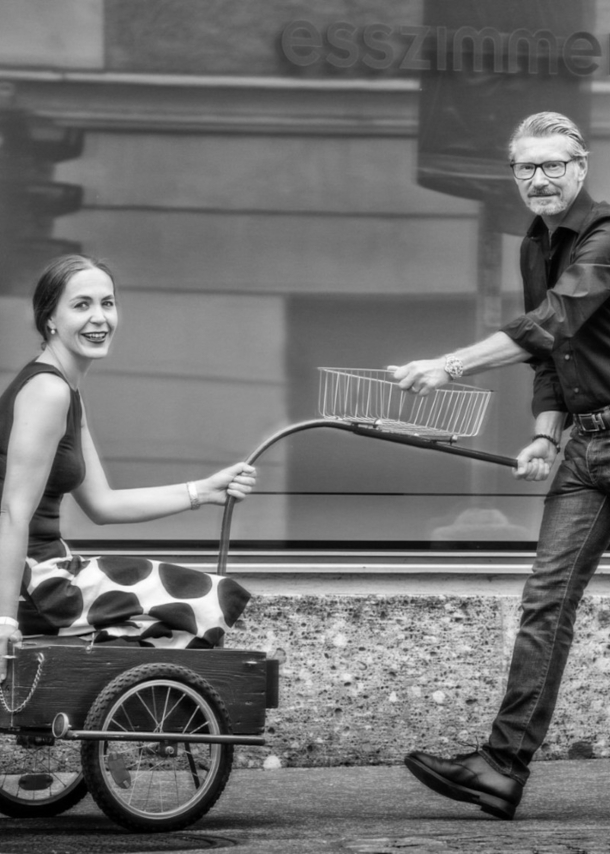 Ein Mann zieht eine lächelnde Frau in einem Fahrradanhänger auf einem Gehweg vor einer Fensterfront.