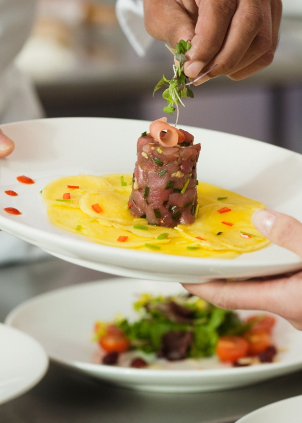 Ein Koch garniert ein Gericht mit Thunfischtartar auf einem weißen Teller in einer Restaurantküche.