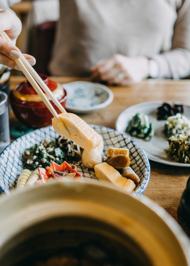 Auf einem Tisch stehen mehrere Schälchen mit verschiedenen asiatischen Gerichten. Eine unkenntliche Person hält mit Stäbchen etwas zu essen hoch.