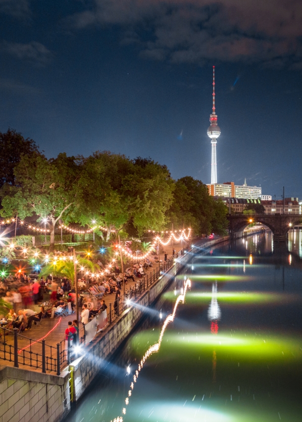 Personen in einer Strandbar am Spreeufer an der Berliner Museumsinsel bei Nacht, im Hintergrund ein beleuchteter Fernsehturm.