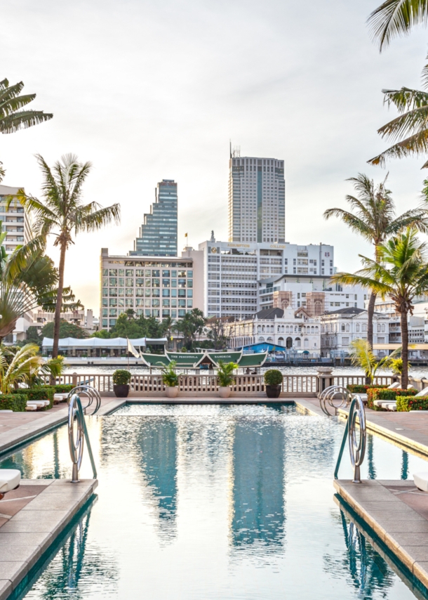 Eine mit Palmen gesäumte Poolanlage im Hotel The Peninsula in Bangkok. Am Wasser stehen Holzliegen mit weißem Bezug, einige befinden sich unter Pagoden-Dächern.