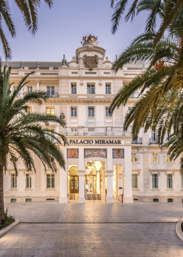 Imposante, helle Frontfassade eines Grandhotels mit Vorplatz, im Vordergrund Begrünung mit Palmen.