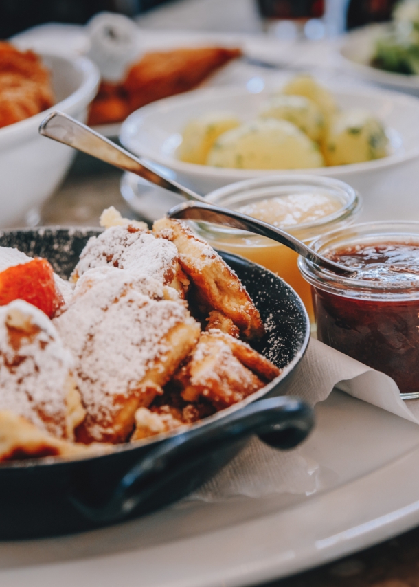 Ein schwarzer Topf mit Kaiserschmarrn auf einem weißen Teller, drumherum kleine Schälchen mit verschiedenen Leckereien.