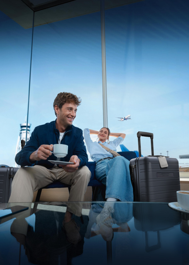 Zwei Personen sitzen nebeneinander in einer Flughafen-Lounge, neben sich jeweils einen Rollkoffer der Marke HORIZN, und trinken einen Kaffee.