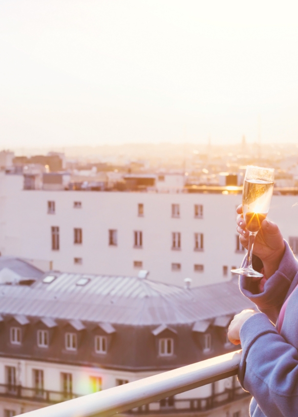 Eine Frau, die bei Sonnenuntergang in Paris auf einem Balkon steht und ein Glas Sekt in der Hand hält.