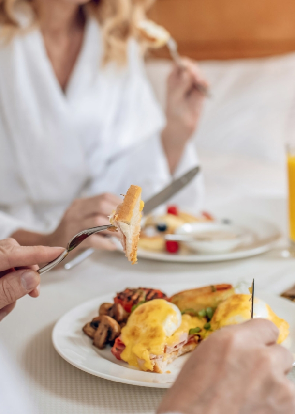 Nahaufnahme von zwei gesichtslosen Personen in weißen Bademänteln, die an einem eingedeckten Tisch in einem Hotelzimmer frühstücken.