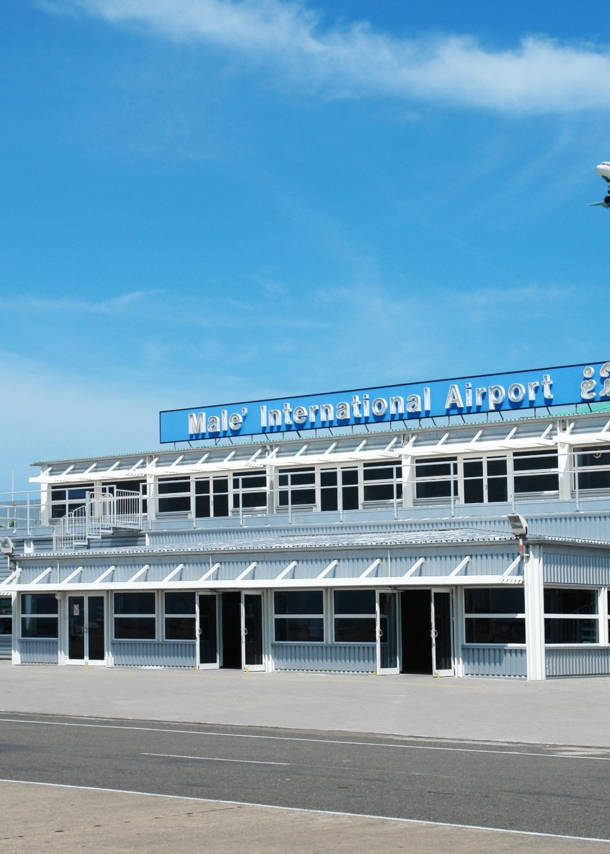 Ein Flugzeug fliegt am blauen Himmel über einen kleines Flughafengebäude mit blauem Kontrollturm.