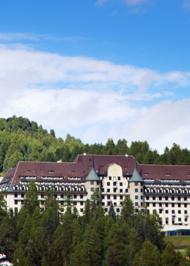 Ein Grandhotel mit zwei Türmen in einem Waldgebiet auf einem Berg, darüber fährt eine Seilbahn.