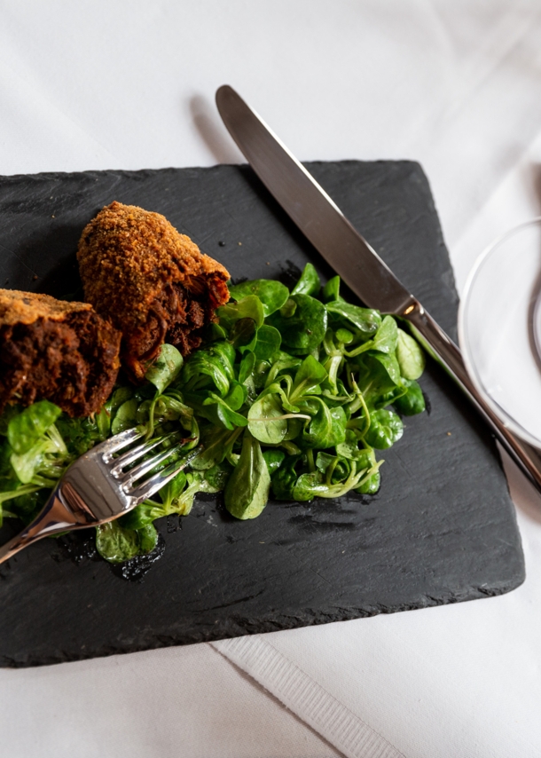 Aufsicht eines panierten Fleischgerichtes an Feldsalat auf einer Schieferplatte auf einem edel gedeckten Tisch, im Vordergrund eine Hand mit einem Glas Weißwein.