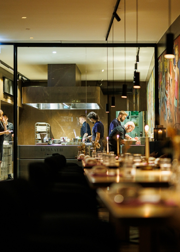 Personen in einer offenen Restaurantküche, davor Speiseraum mit eingedeckten Tischen bei Kerzenschein.