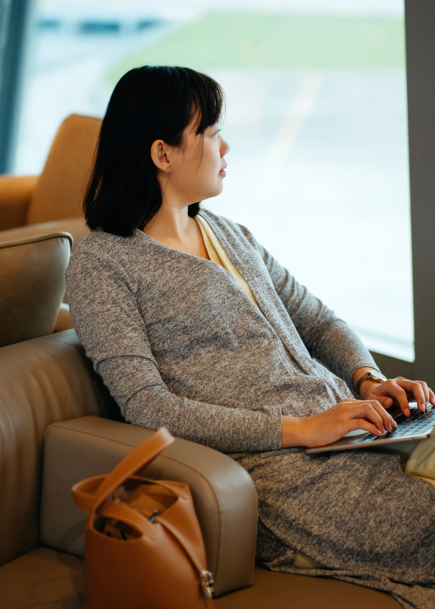 Eine Frau sitzt mit Laptop in einem Ledersessel am Fenster einer Flughafen-Lounge und schaut aufs Rollfeld.