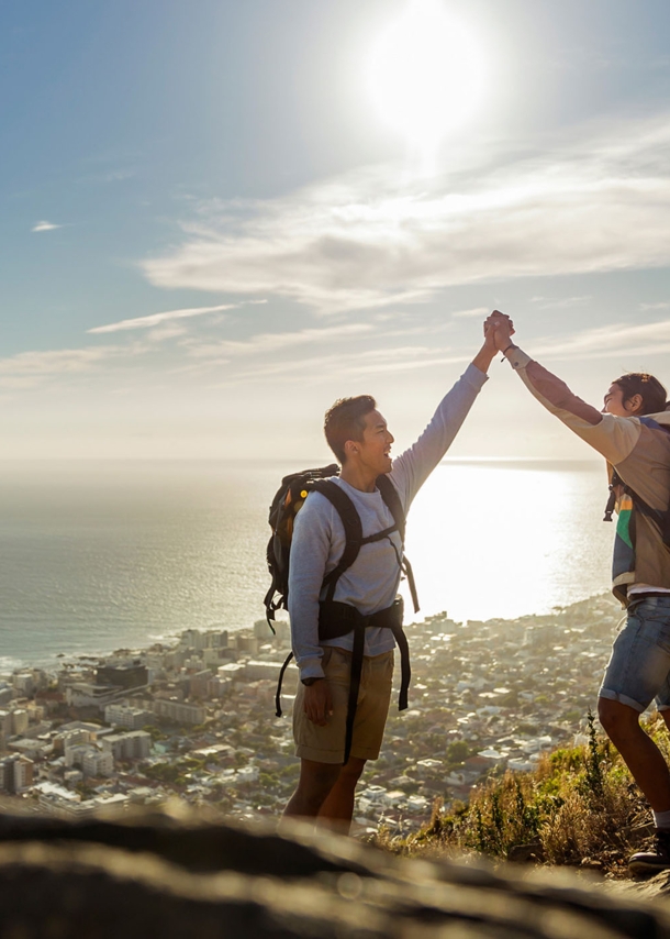 Zwei Wanderer mit Rucksäcken geben sich ein High Five auf einer Bergspitze, im Hintergrund eine Großstadt am Meer.