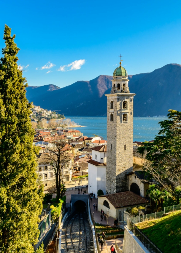 Stadtpanorama von Merano mit Zypressen und Glockenturm an einem See vor Bergpanorama.