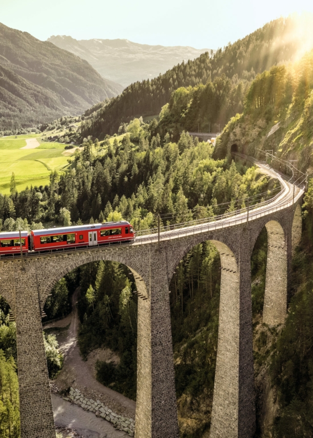 Ein roter Zug fährt auf einem Viadukt durch eine grüne Berglandschaft.