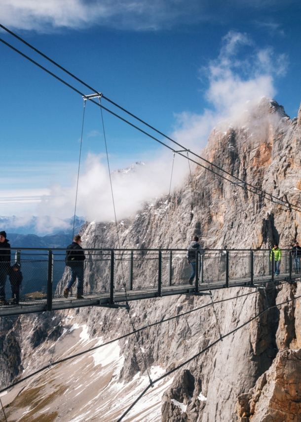 Personen auf einer Hängebrücke vor einer Felswand im Gebirge.