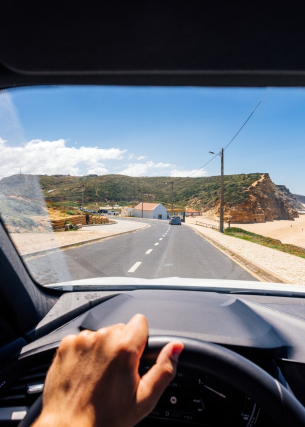 Blick über ein Lenkrad durch die Frontscheibe eines fahrenden Autos auf einer Straße an einer Küste mit Sandstrand bei Sonnenschein.