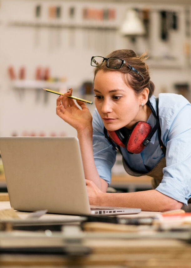 Foto einer Handwerkerin, wie sie an ihrer Werkbank steht und auf den vor ihr geöffneten Laptop blickt.