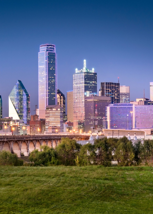 Skyline von Dallas mit Wolkenkratzern hinter einer Grünfläche mit Brückenbauten bei Abenddämmerung.