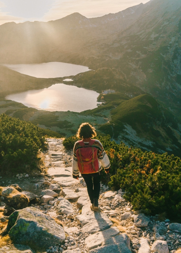 Rückansicht einer Frau im Pullover mit Rucksack auf einem Wanderweg im grünen Gebirge vor zwei Bergseen im Gegenlicht.