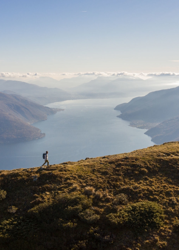 Panoramaaufnahme von einem Mann mit Rucksack auf einem Berg und dem Lago Maggiore im Hintergrund.