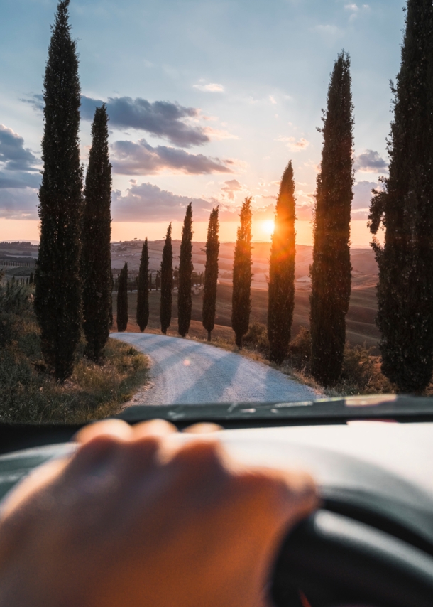 Blick auf eine mit Zypressen gesäumte Landstraße in Hügellandschaft bei Sonnenuntergang durch die Frontscheibe eines Autos, im Vordergrund eine Hand am Lenkrad.