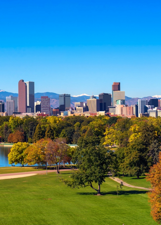 Skyline von Denver unter blauem Himmel vor Gebirgszug, im Vordergrund eine Parkanlage mit Teich und grünen Bäumen.