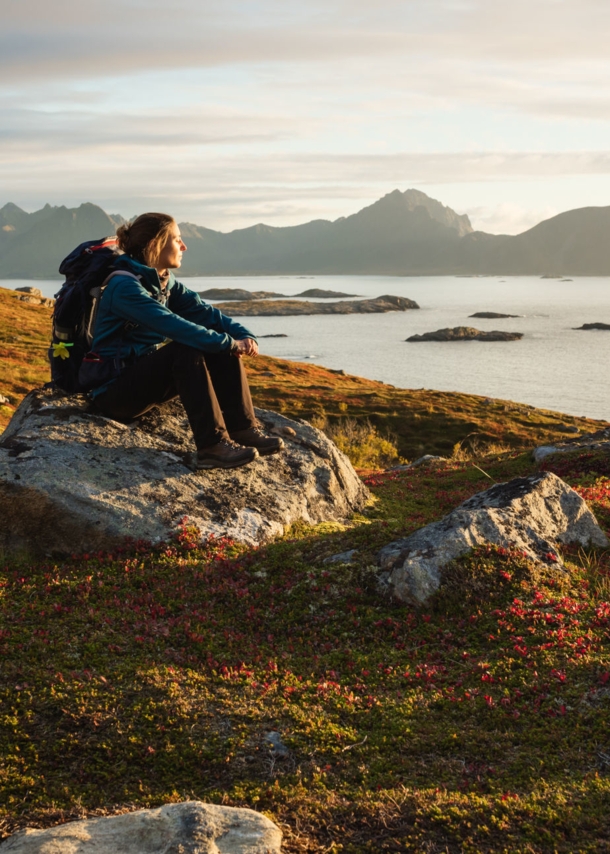 Eine Frau mit Wanderausrüstung, die bei Sonnenuntergang auf einem großen Stein sitzt und aufs Wasser blickt, im Hintergrund Berge.